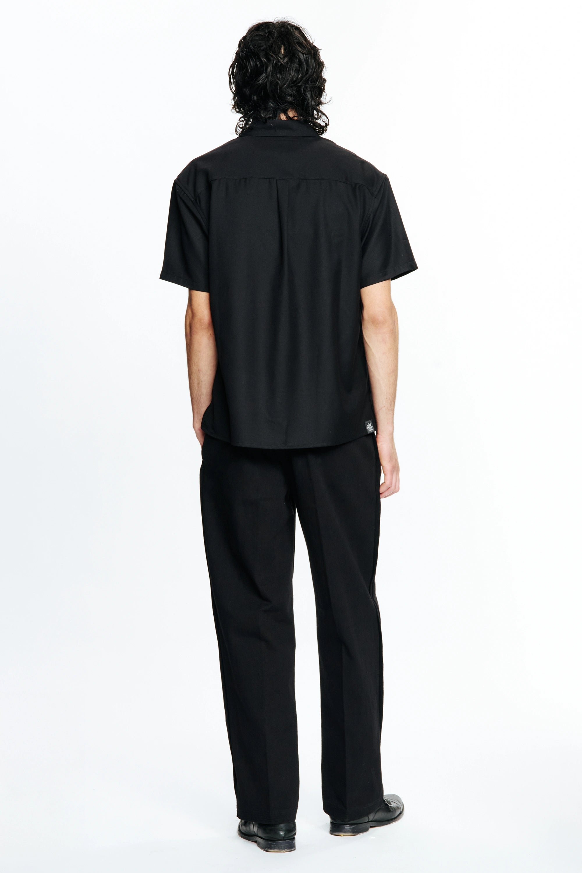 Person with their back turned wearing a Black Hold Short Sleeve Shirt and dark trousers on a white background