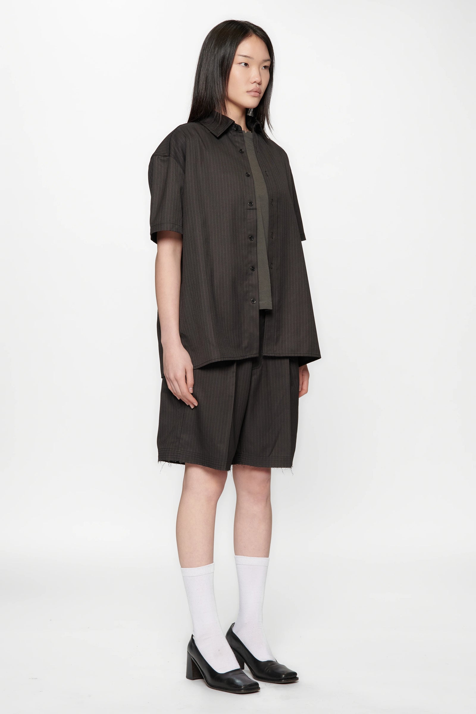 Female model facing to the side wearing a Classic Pinstripe Hold Short Sleeve Shirt and dark shorts with black shoes and white socks