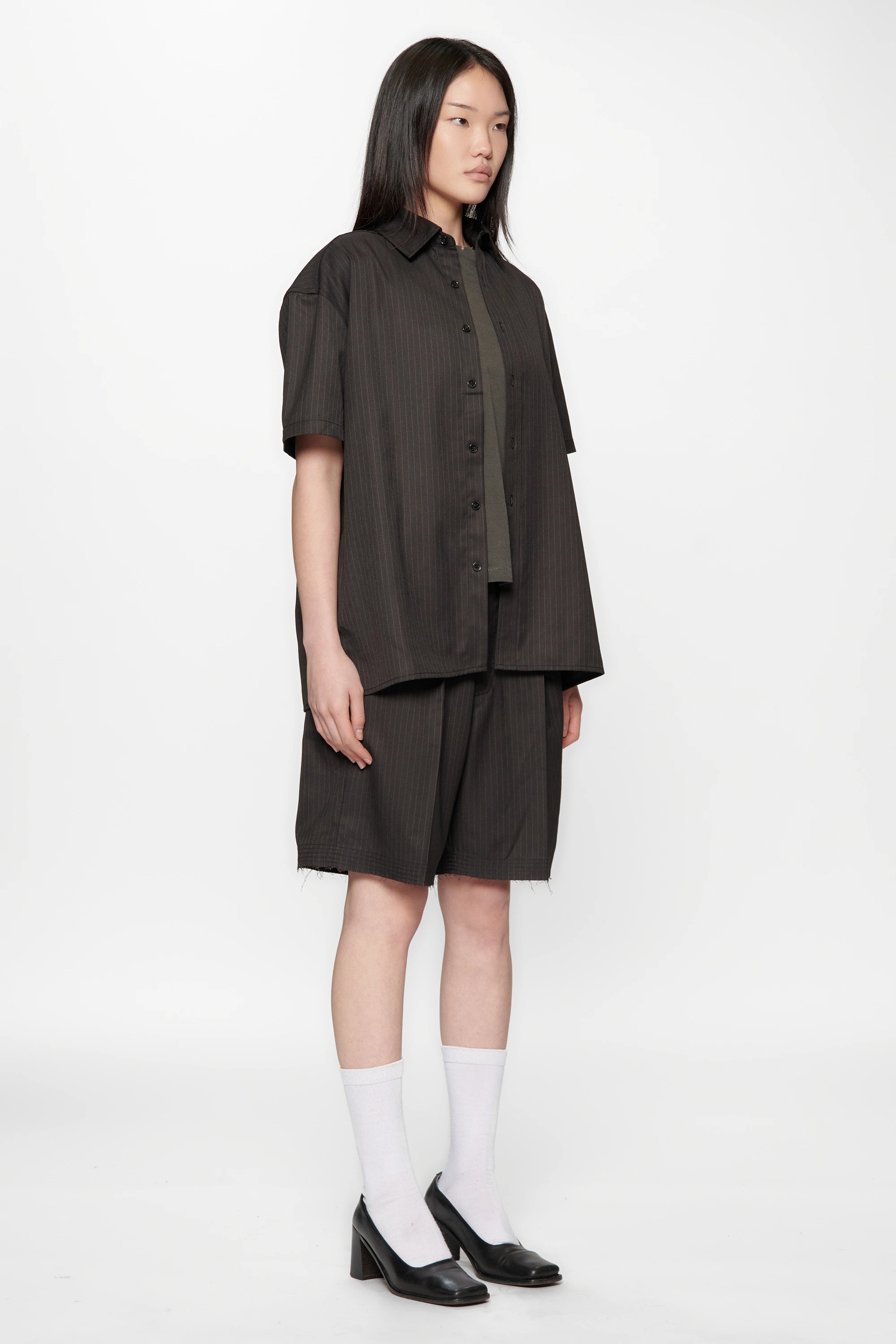 Female model facing to the side wearing a Classic Pinstripe Hold Short Sleeve Shirt and dark shorts with black shoes and white socks