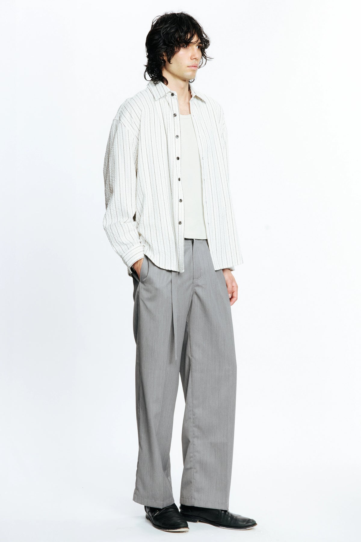 Man looking to the side with one hand resting in a pocket wearing a white  long-sleeve striped shirt and grey pants on a white background