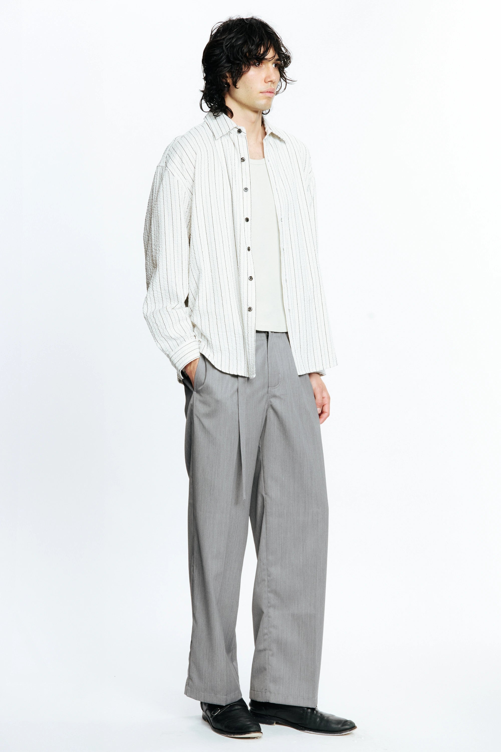 Man looking to the side with one hand resting in a pocket wearing a white  long-sleeve striped shirt and grey pants on a white background