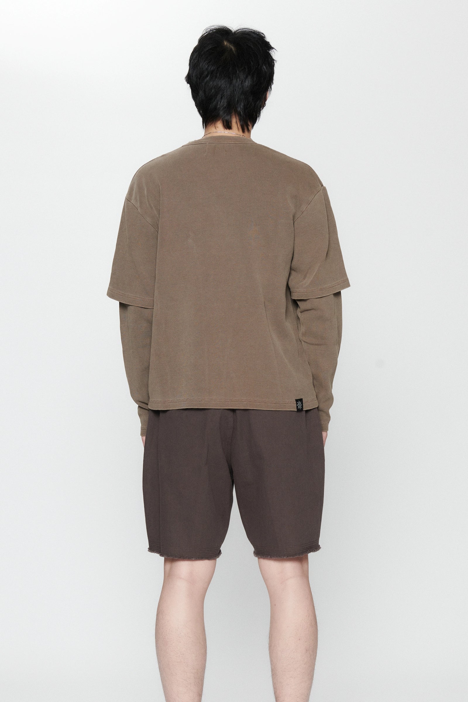 A person standing facing a white background weraing a pair of pleated shorts in a dark grey color and a long-sleeve shirt.