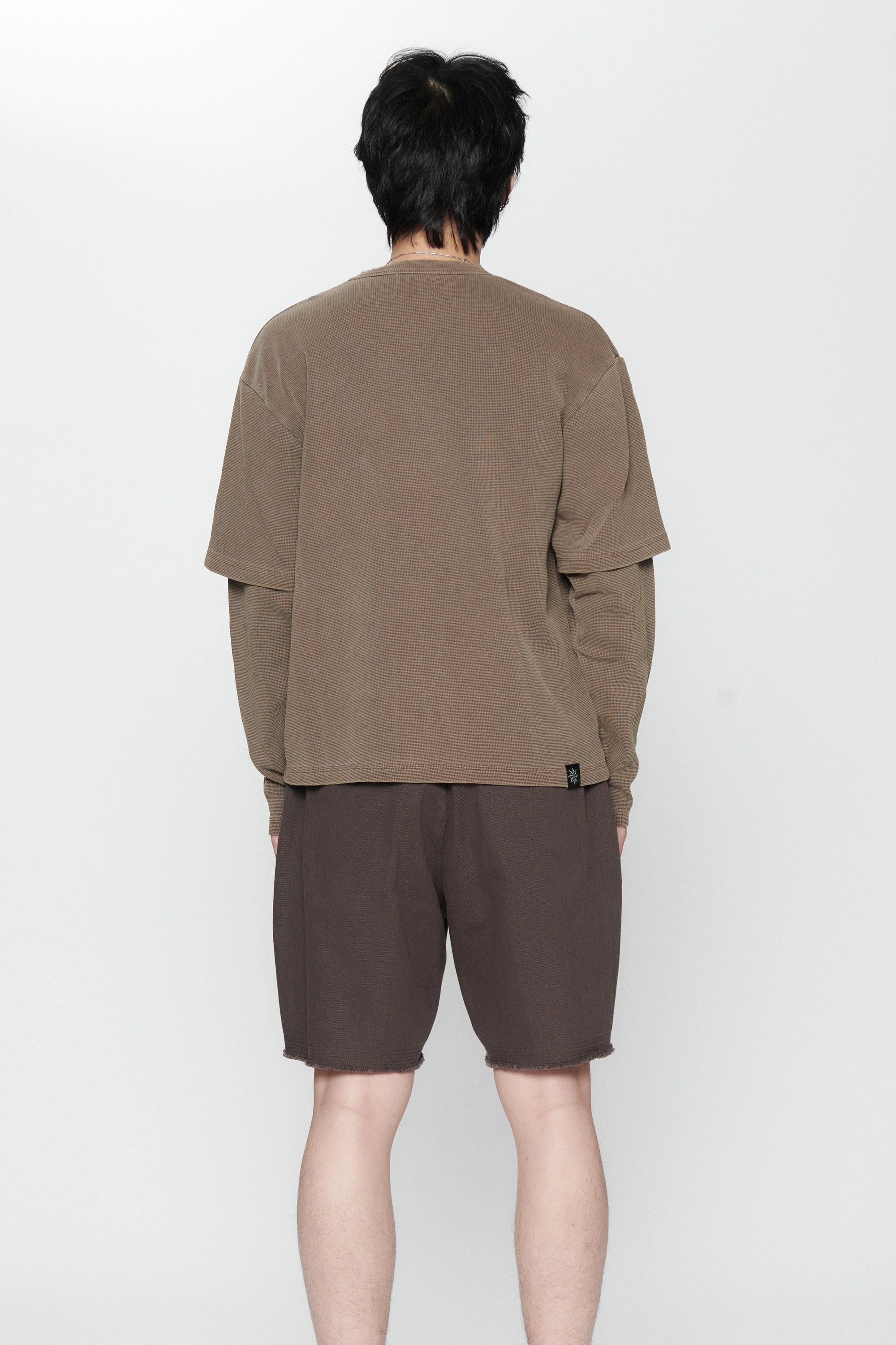 A person standing facing a white background weraing a pair of pleated shorts in a dark grey color and a long-sleeve shirt.