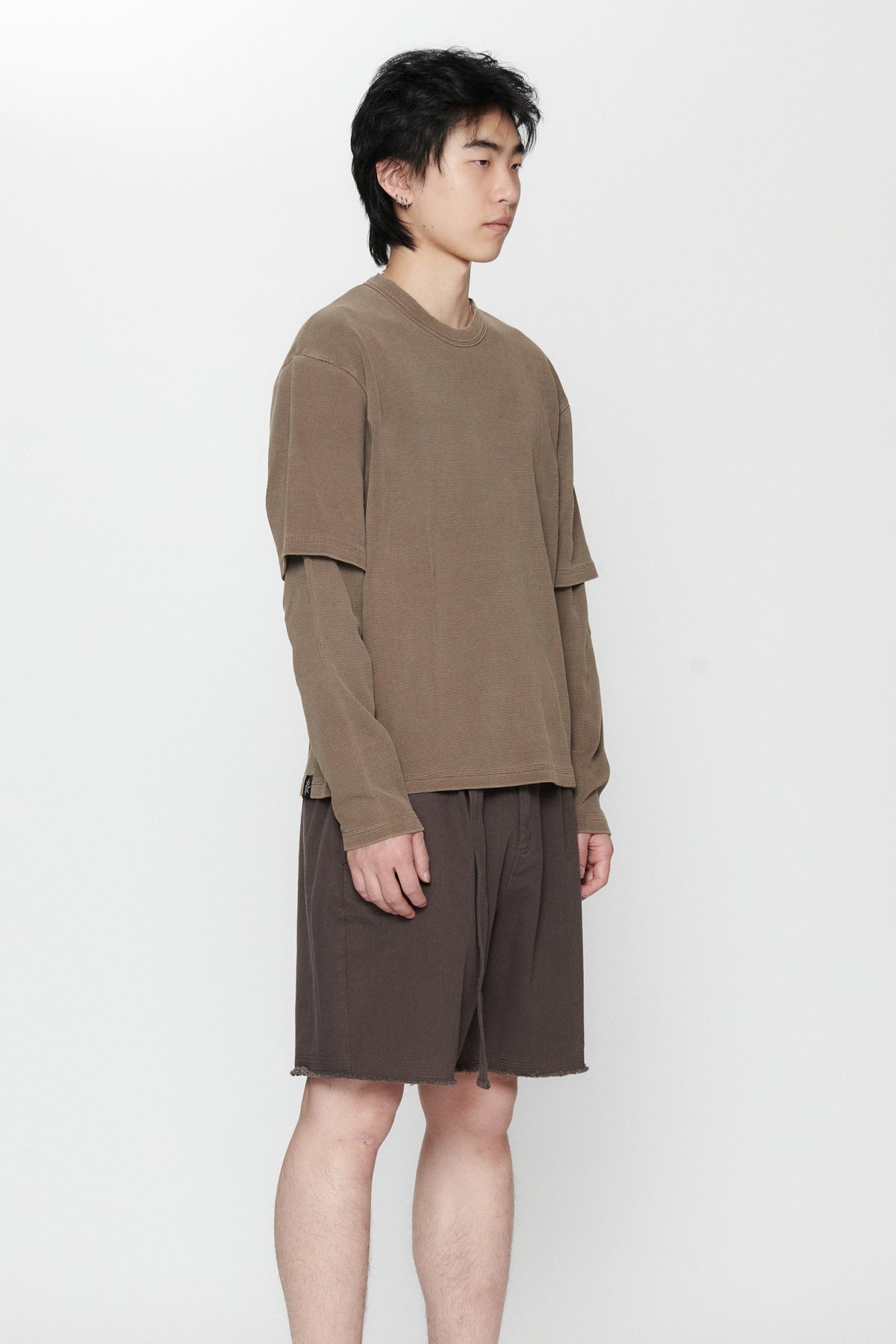 A person standing against a white background facing to the side wearing a dark grey pleated long-sleeve top and matching shorts.