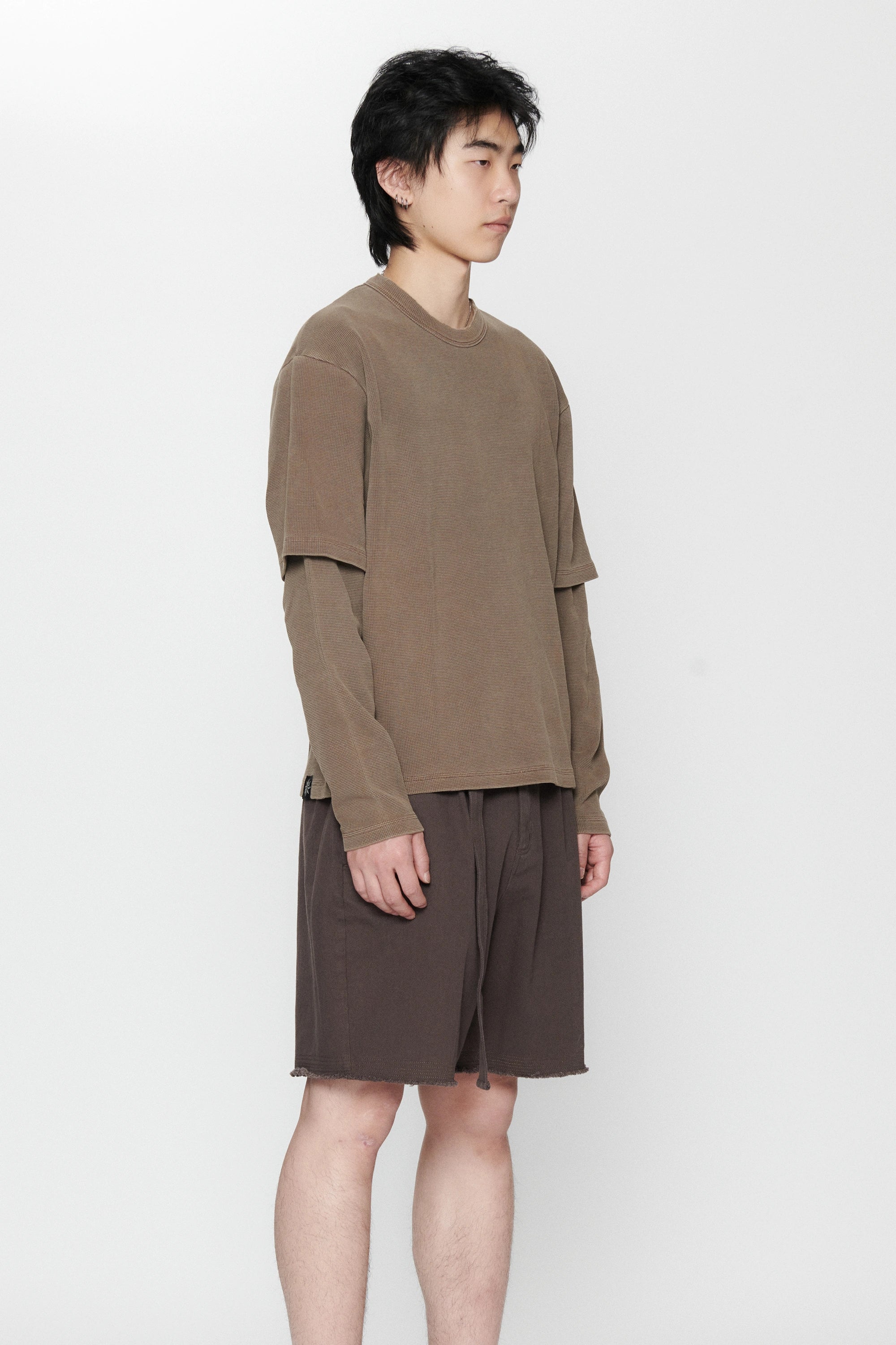A person standing against a white background facing to the side wearing a dark grey pleated long-sleeve top and matching shorts.