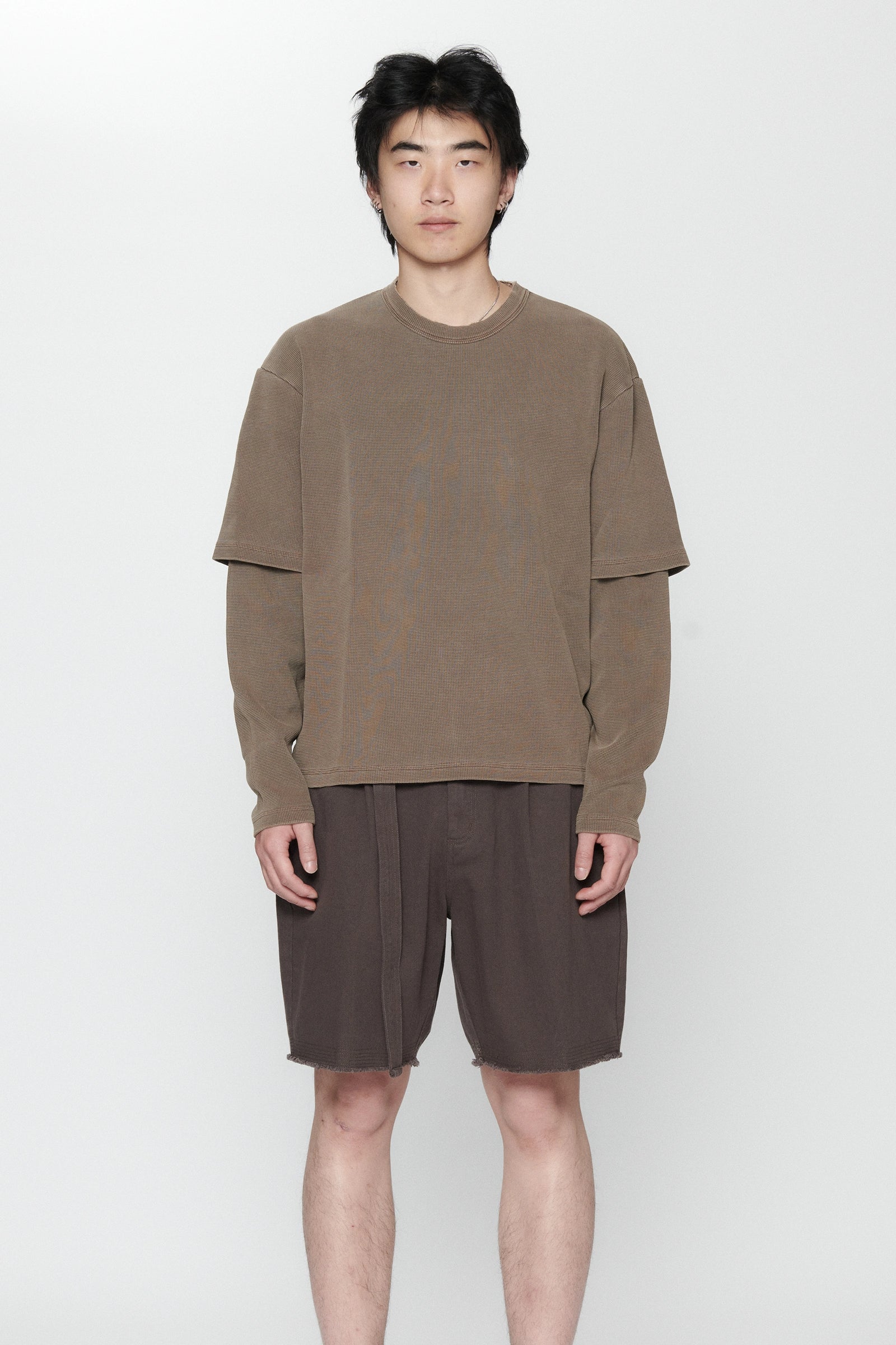 A person standing against a white background wearing a dark grey pleated long-sleeve top and matching shorts.