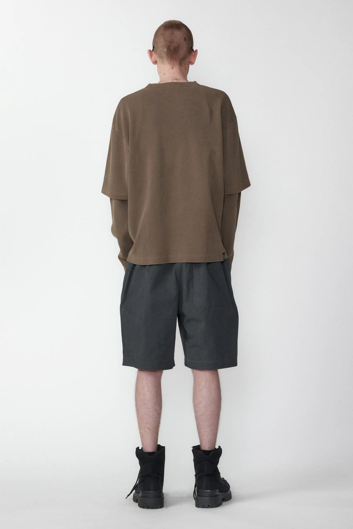 A person with their back turned standing against a white background wearing relaxed fit charcoal grey shorts and a beige long-sleeve shirt