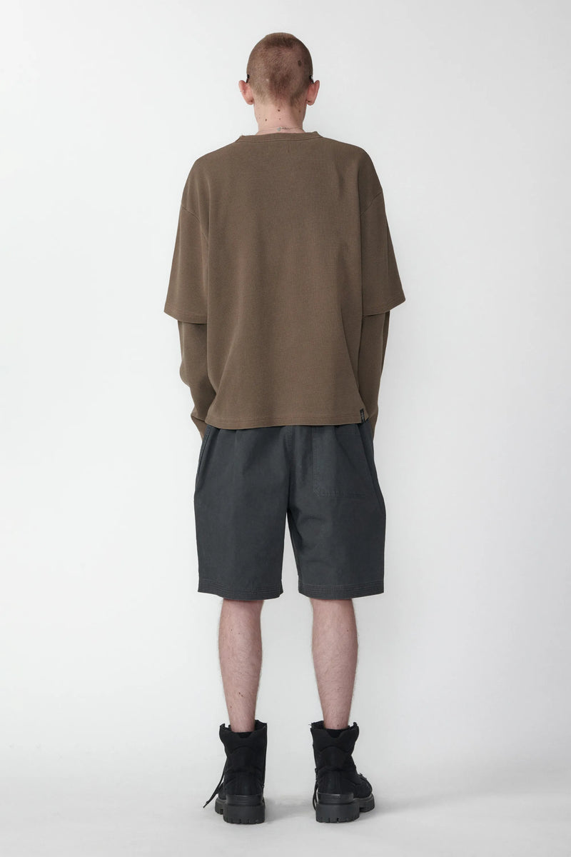A person with their back turned standing against a white background wearing relaxed fit charcoal grey shorts and a beige long-sleeve shirt