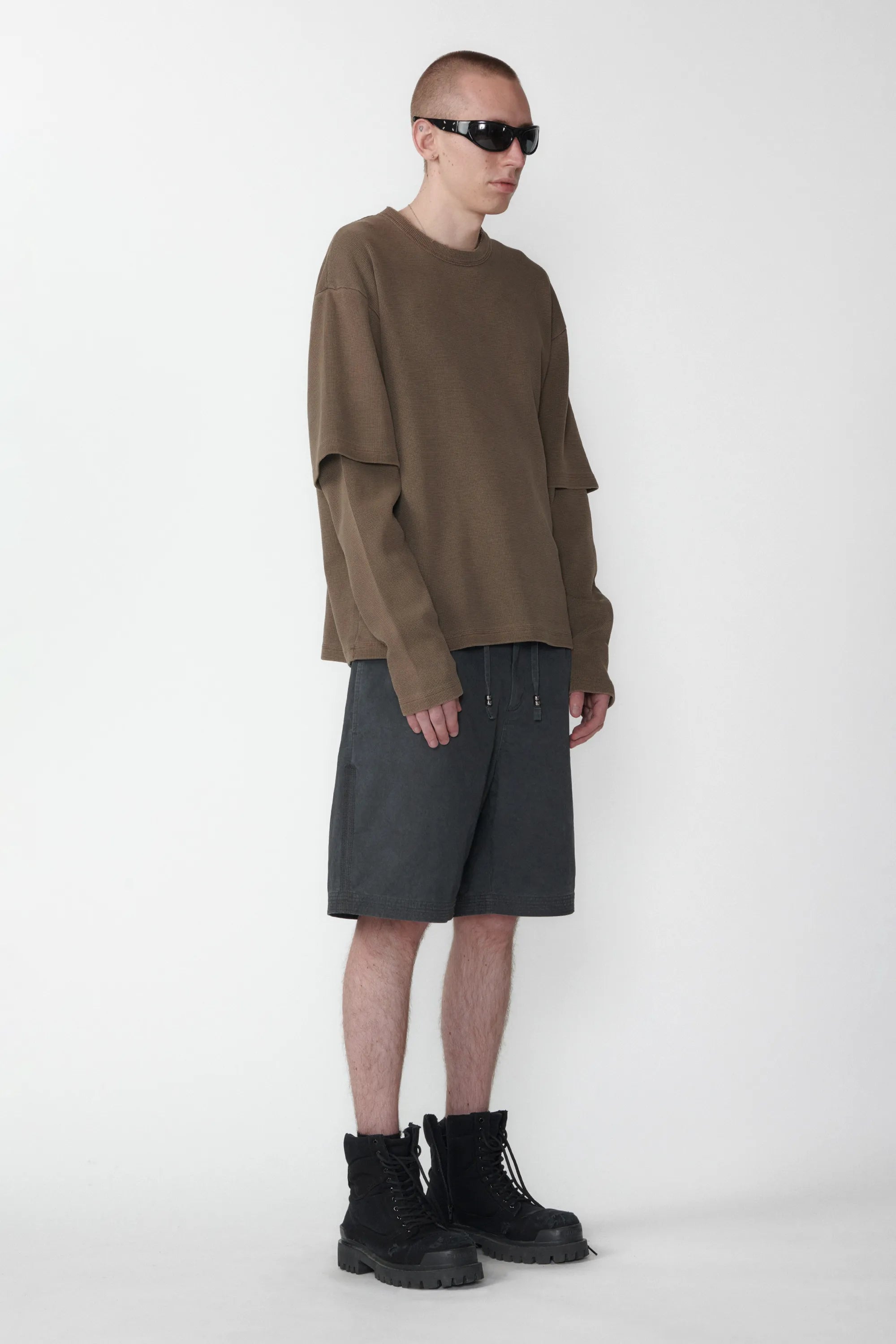 A person standing sideways wearing relaxed fit charcoal grey shorts with an elastic waistband and a beige long-sleeve shirt on a white background