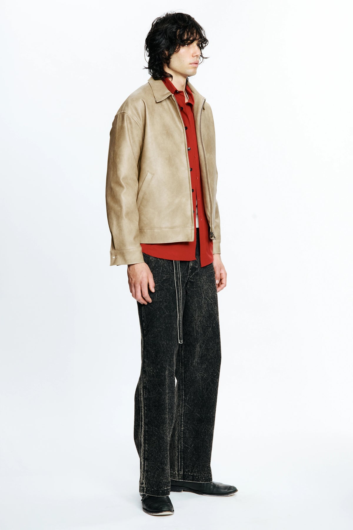 Man facing to one side wearing a beige leather jacket over a red shirt with black pants on a white background