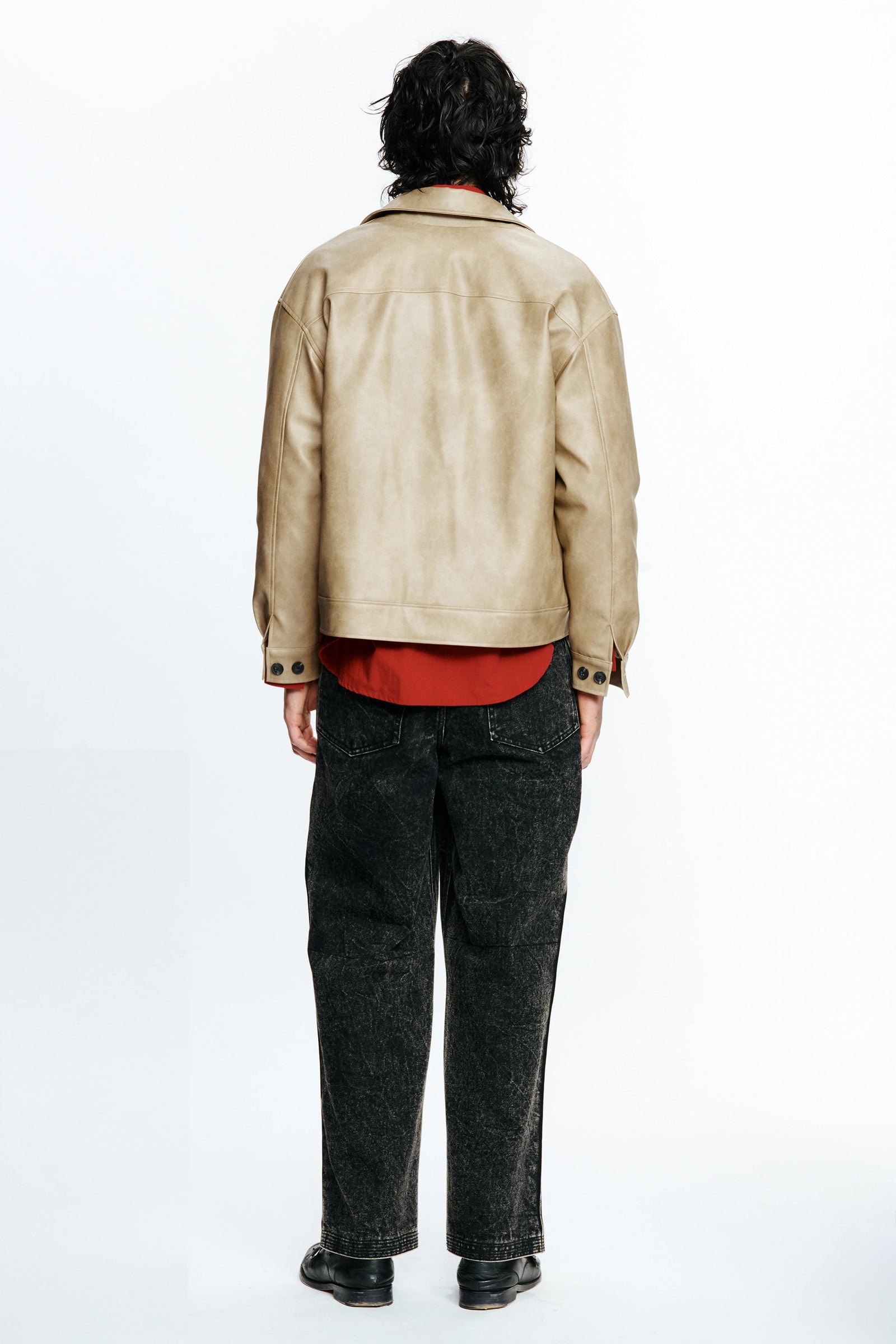Person with their back turned wearing a beige leather jacket, red shirt and dark jeans on a white background