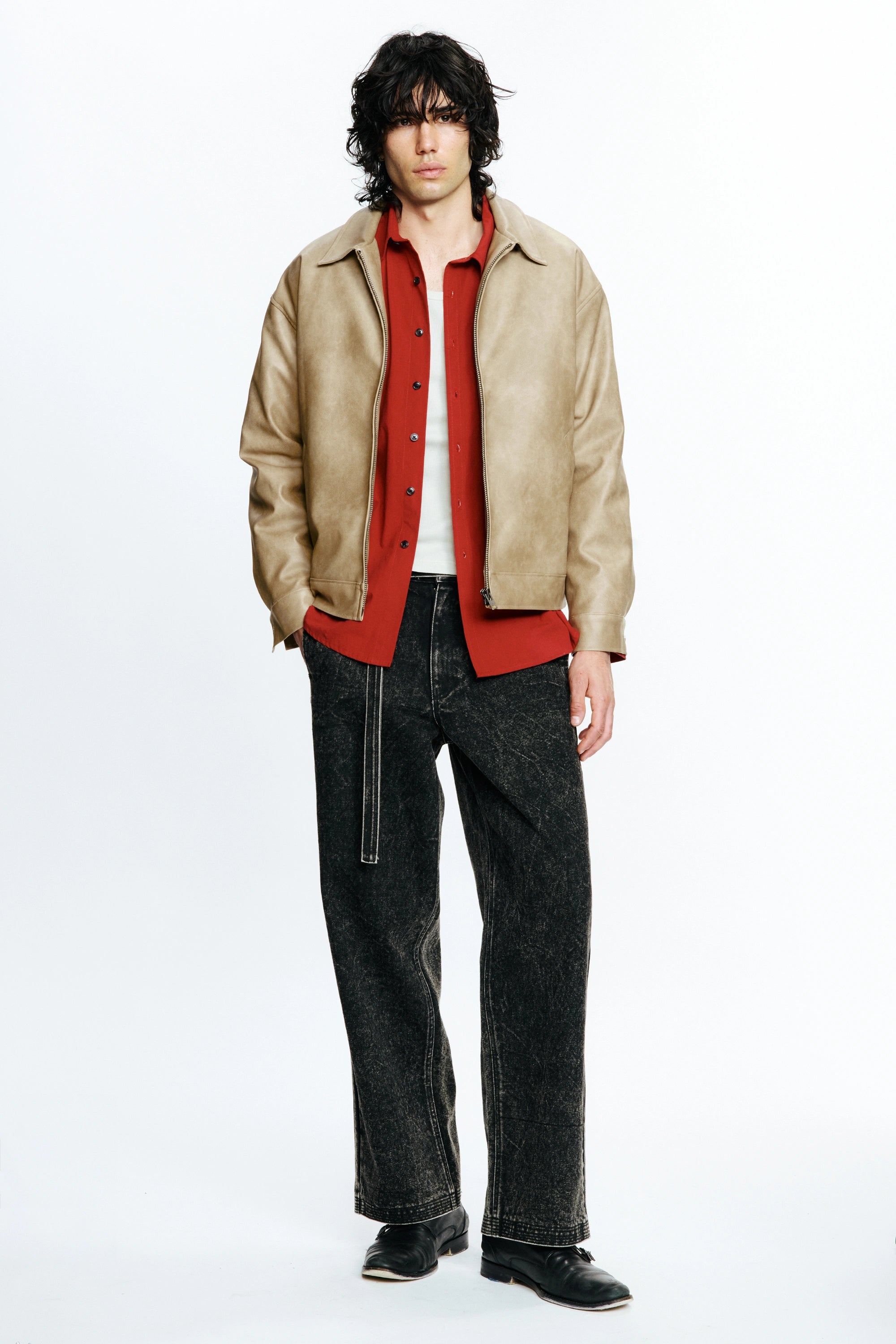 Man wearing a beige leather jacket over a red shirt with black pants on a white background