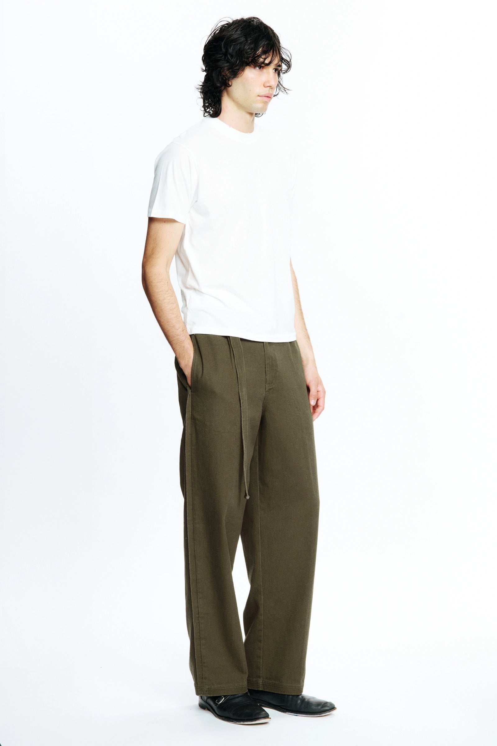 A person standing against a white background facing off to the side with one hand in their pocket wearing a white t-shirt and pleated work pants in a green colour.