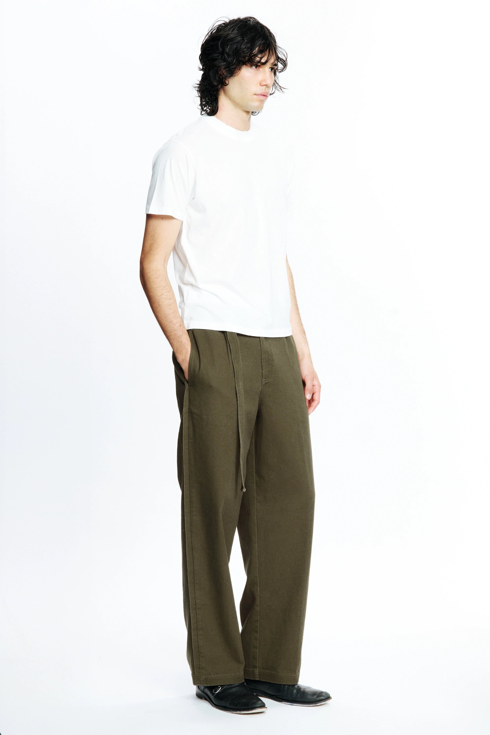 A person standing against a white background facing off to the side with one hand in their pocket wearing a white t-shirt and pleated work pants in a green colour.