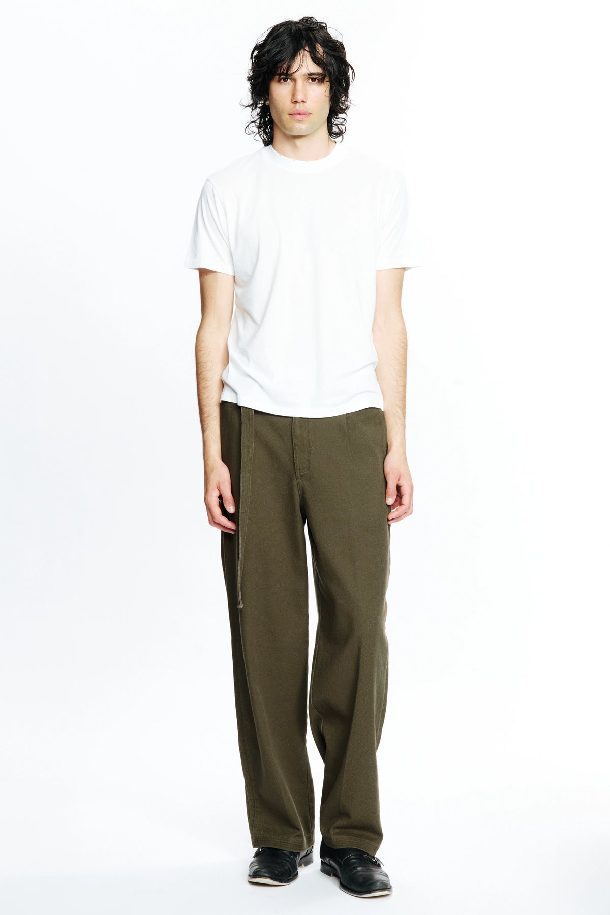 A person standing against a white background, wearing a white t-shirt and pleated work pants in a green colour.