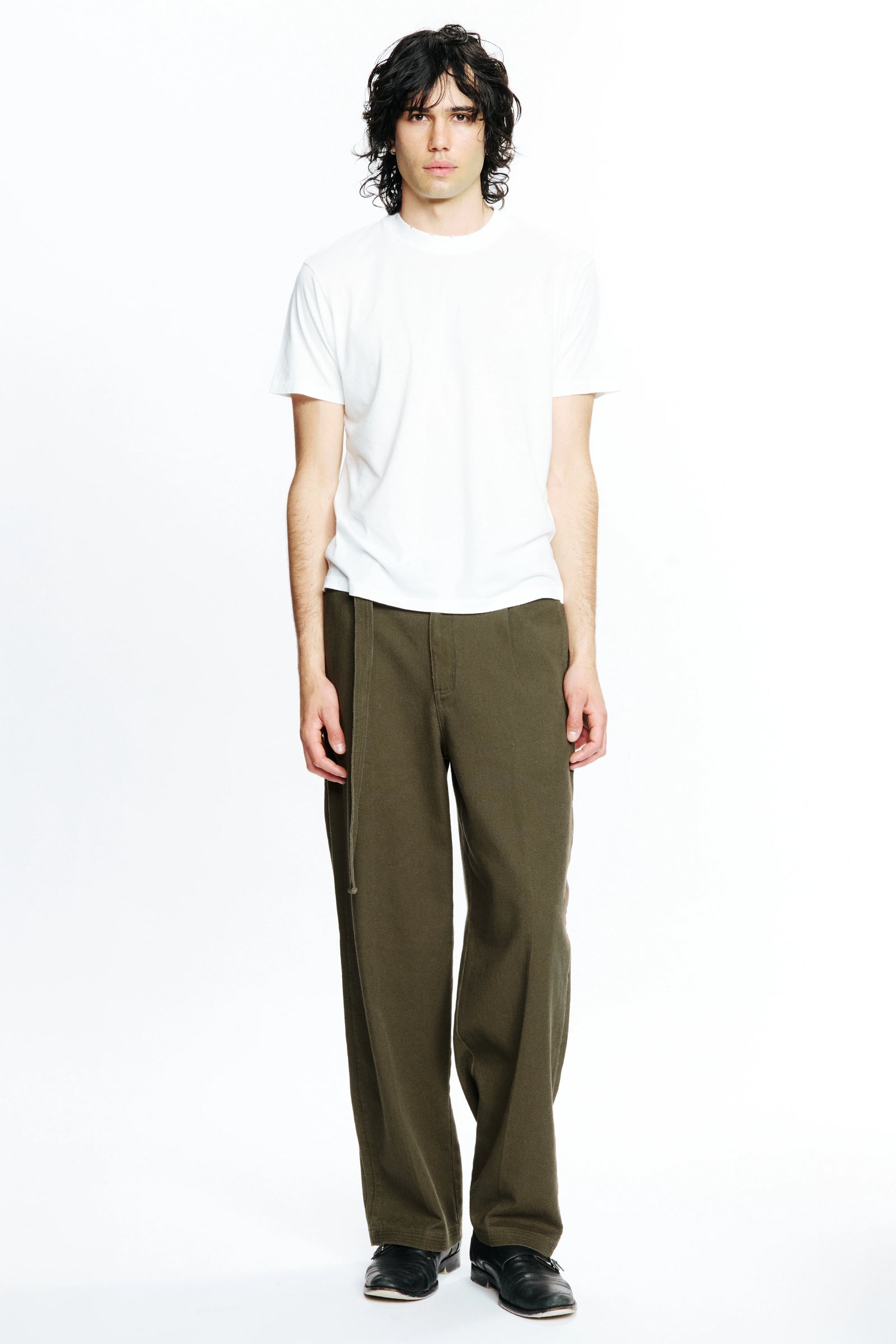 A person standing against a white background, wearing a white t-shirt and pleated work pants in a green colour.