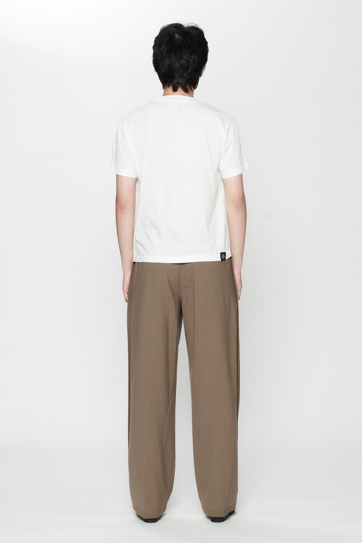 View from behind of a person standing against a white background wearing a white t-shirt and pleated work pants in a brown colour.
