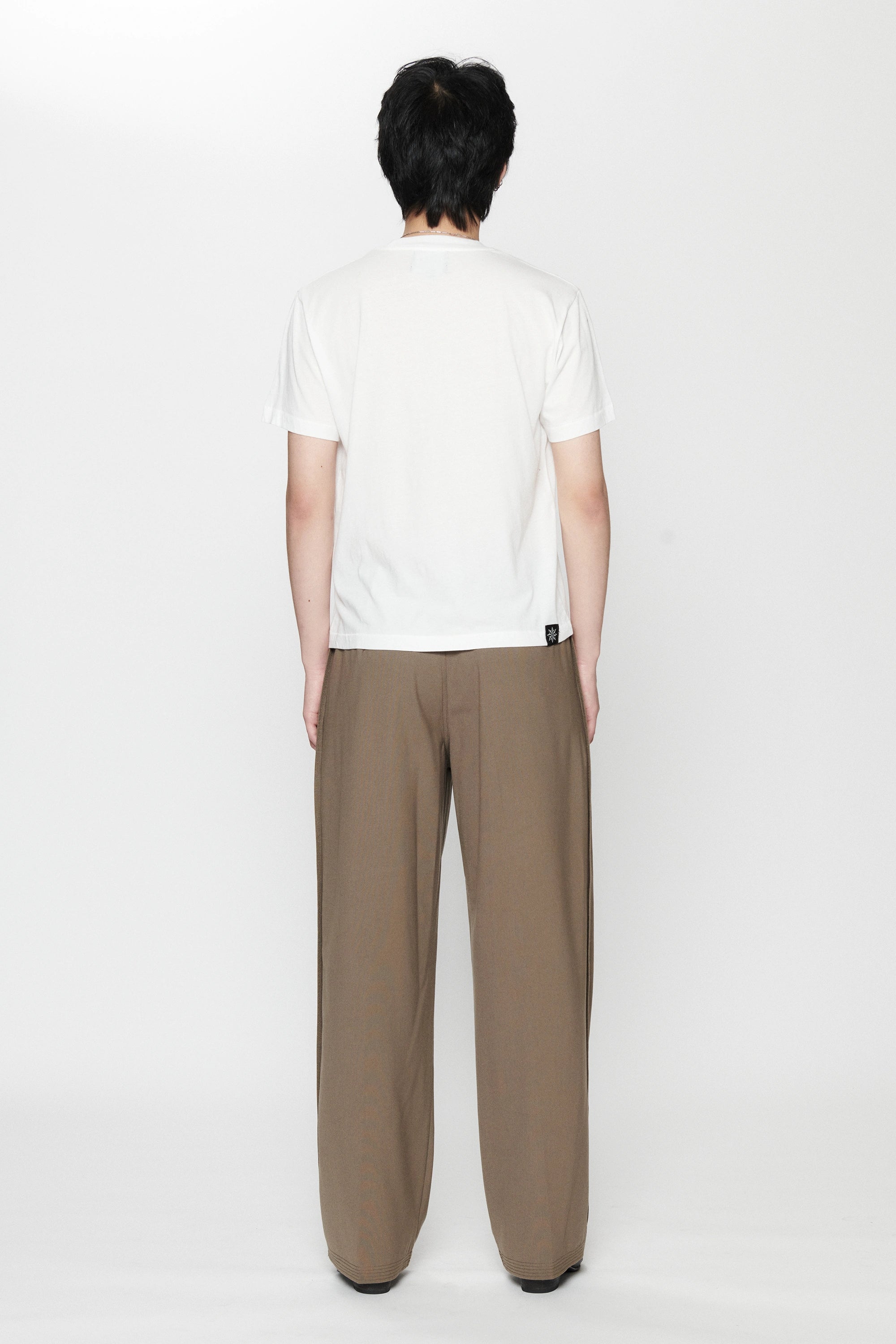 View from behind of a person standing against a white background wearing a white t-shirt and pleated work pants in a brown colour.