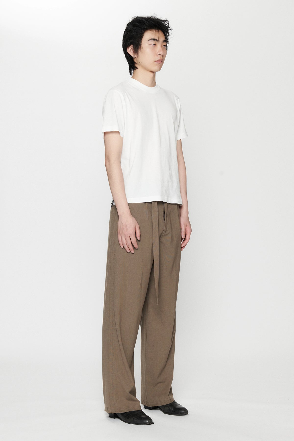 A person standing sideways against a white background wearing a white t-shirt and pleated work pants in a brown colour.