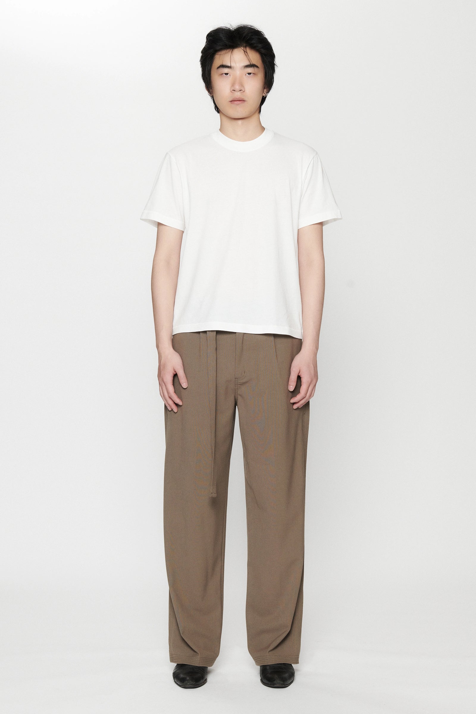 A person standing against a white background, wearing a white t-shirt and pleated work pants in a brown colour.