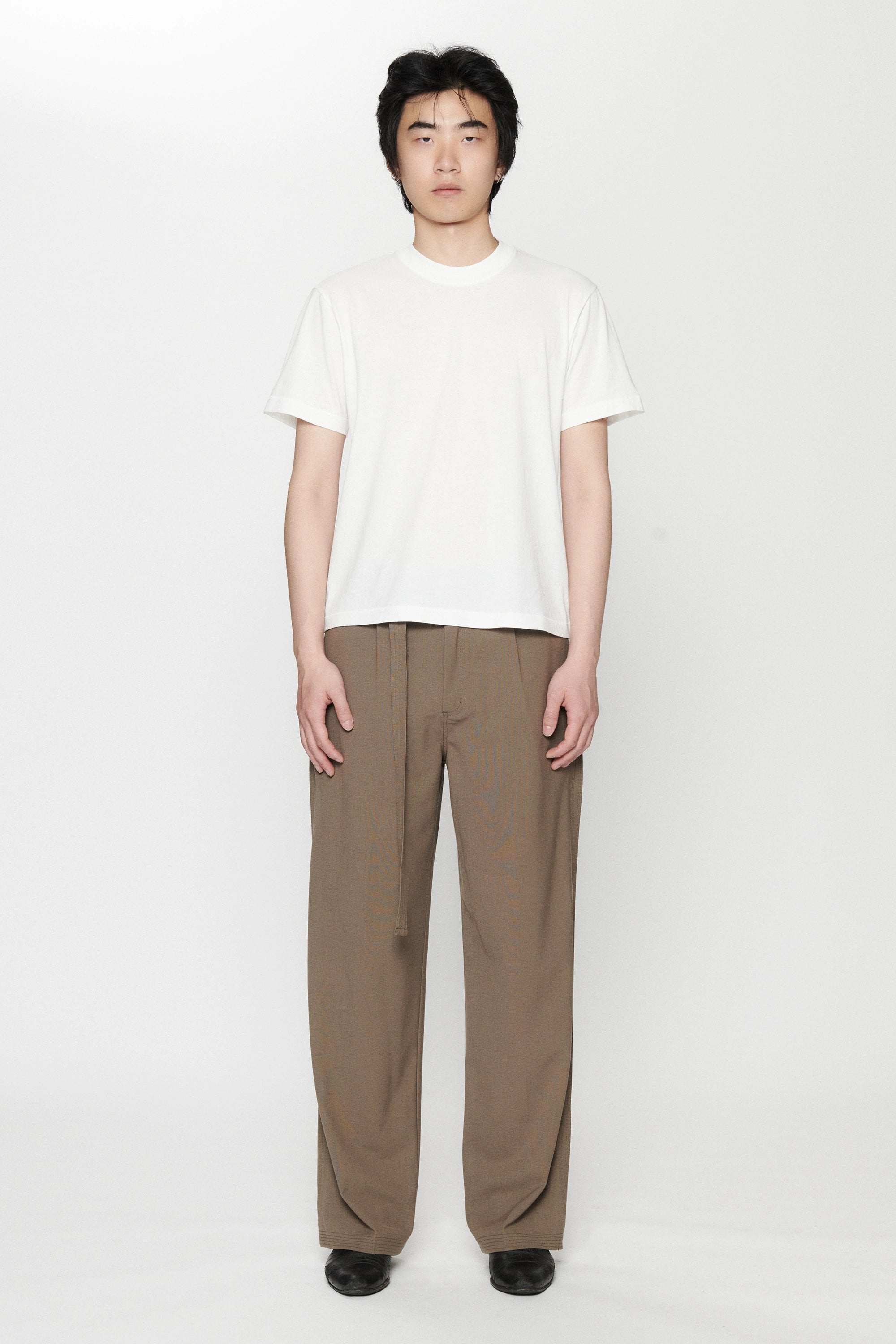 A person standing against a white background, wearing a white t-shirt and pleated work pants in a brown colour.