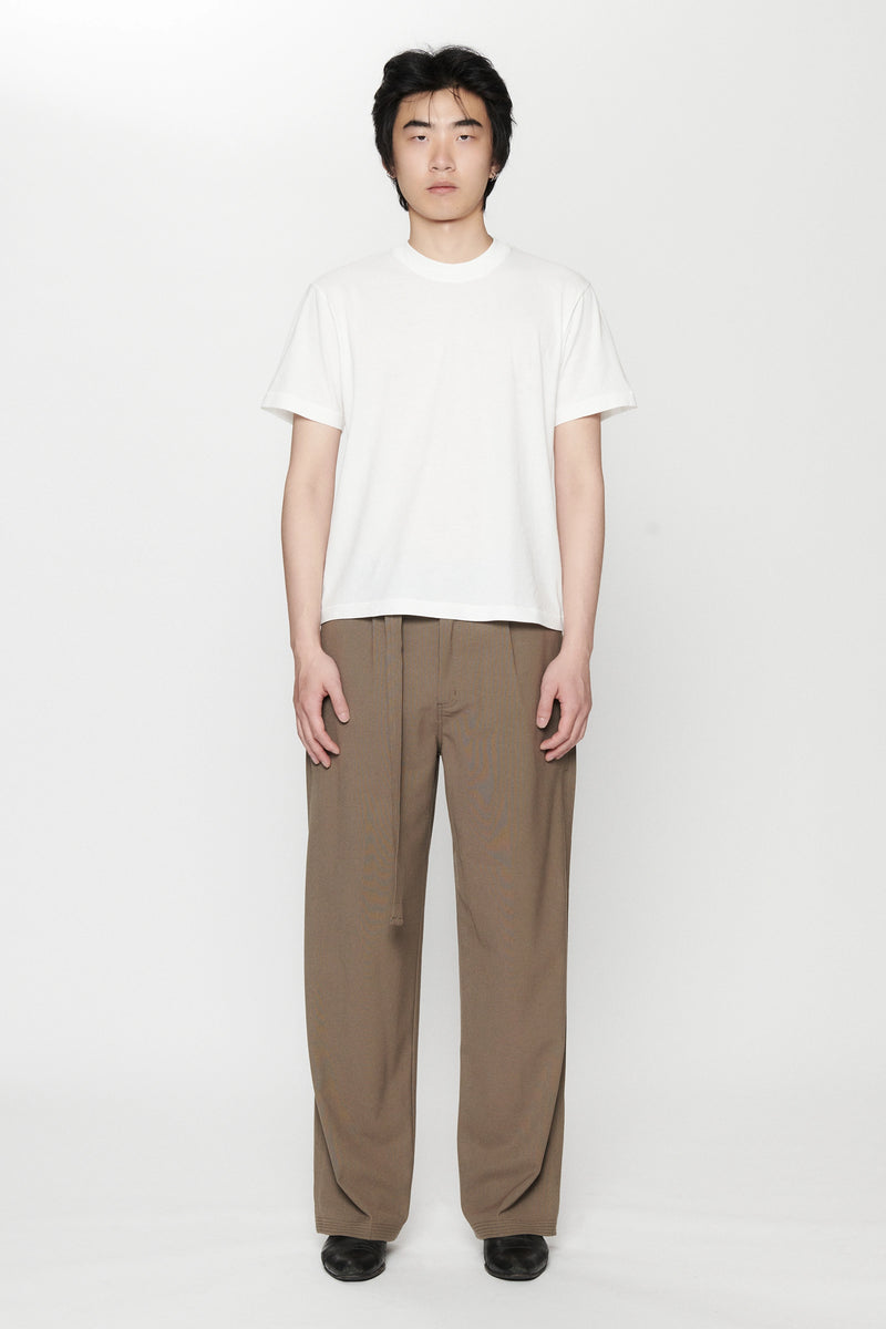 A person standing against a white background, wearing a white t-shirt and pleated work pants in a brown colour.